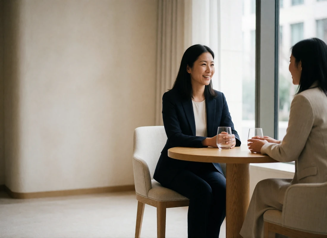 Two Asian women in business-casual attire chatting warmly over coffee in a bright hotel lounge