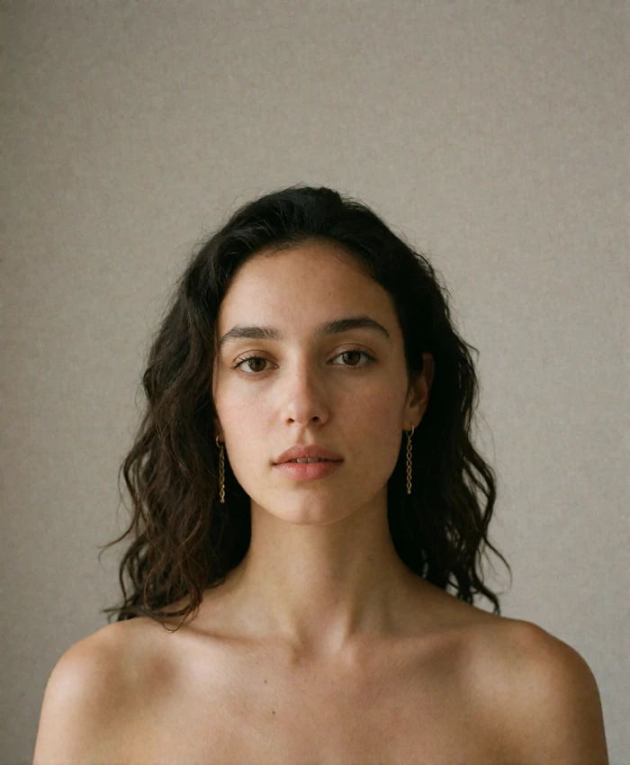 Composed Jordanian woman with wavy dark hair and gold earrings photographed against a warm-grey backdrop
